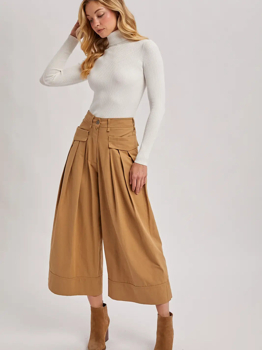Woman wearing a white turtleneck and tan high-waisted pants on a white background