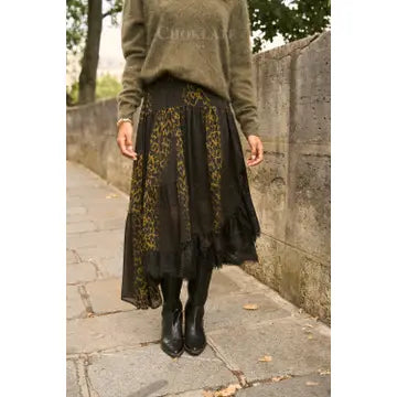 A person standing outdoors on a stone walkway wearing a long olive green sweater and a black midi skirt with yellow floral patterns, paired with black ankle boots.