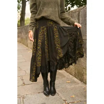 A person outdoors wearing a long black skirt with olive green and black floral lace panels, paired with a green sweater and black knee-high boots, holding out one side of the skirt to show its layered ruffle design.