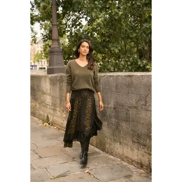A woman walking outdoors on a stone pathway beside a wall and trees, wearing an olive green long-sleeve sweater and a black midi skirt with gold floral embroidery.