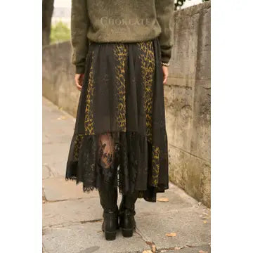 A person standing outdoors on a stone walkway wearing a long black skirt with yellow floral patterns and sheer black lace ruffles, paired with black boots and an olive green sweater.