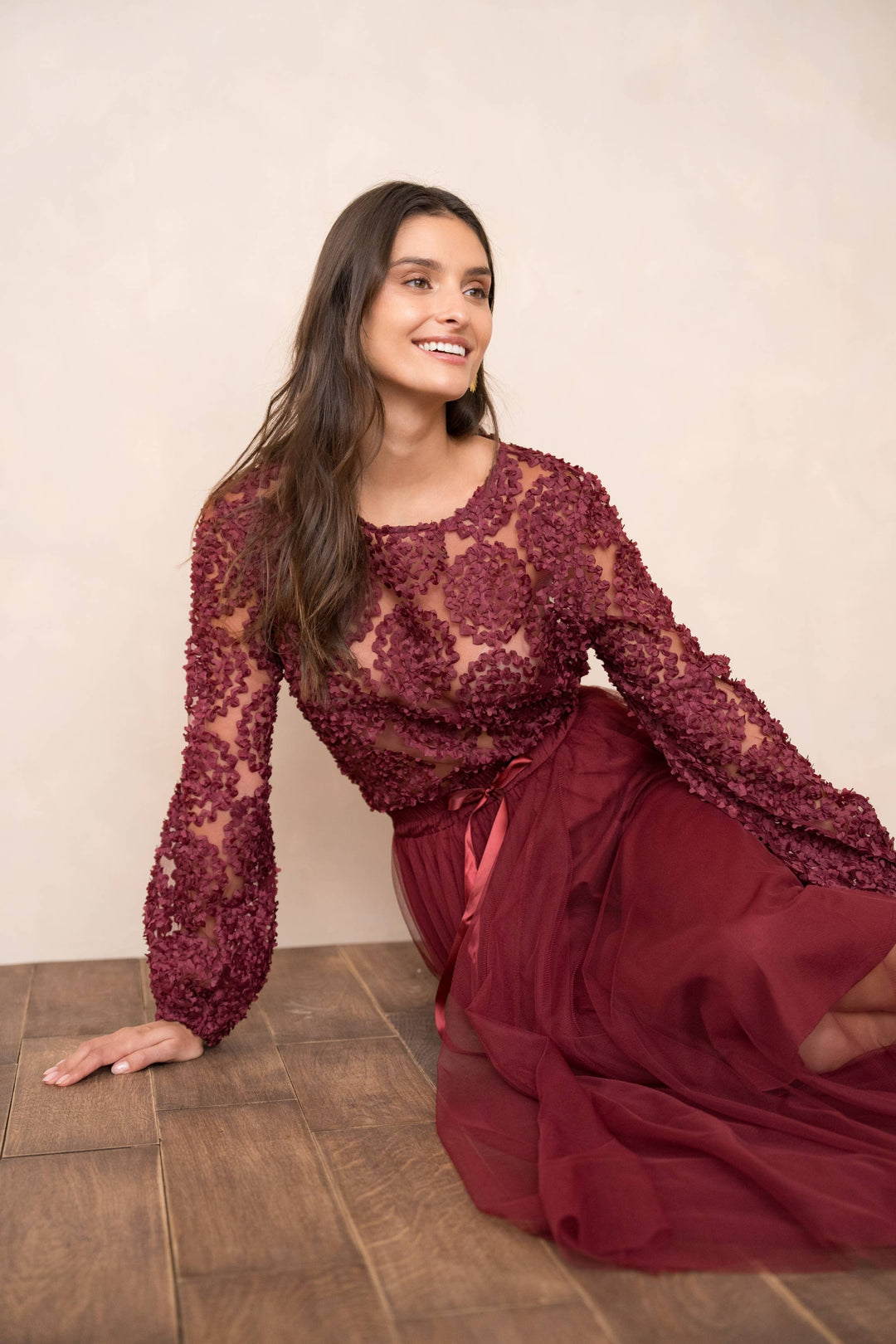 Woman wearing a burgundy lace top and skirt sitting on a wooden floor.