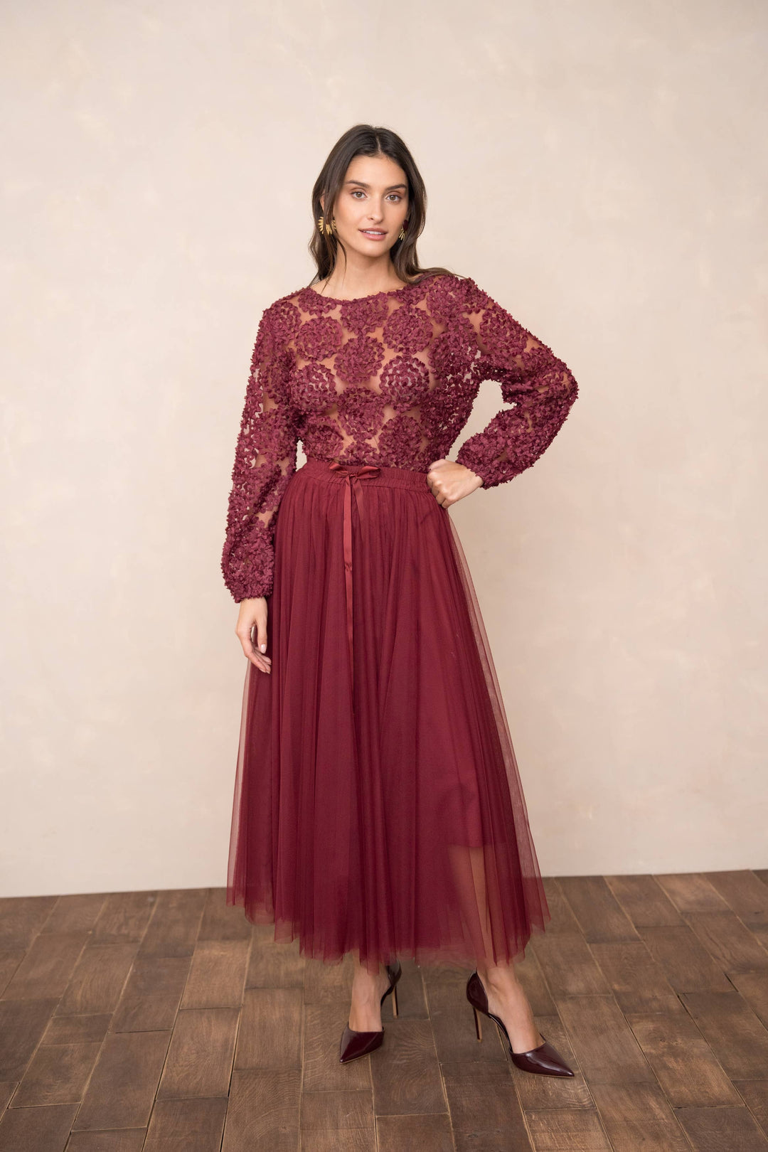 Woman wearing a burgundy lace top and tulle skirt on a wooden floor with a beige wall background