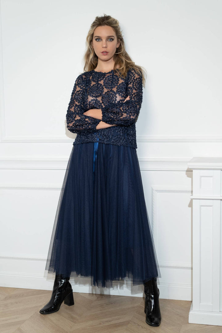 Woman wearing a navy blue lace top and pleated skirt with black boots against a white wall.