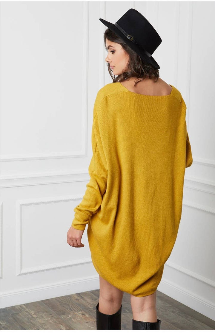 Woman wearing a yellow sweater dress and black hat against a white wall.