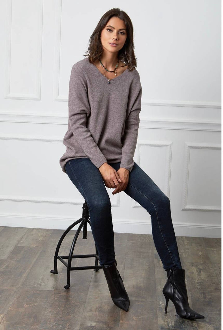 Woman sitting on a stool wearing a gray sweater, dark jeans, and black boots in a room with white paneled walls.