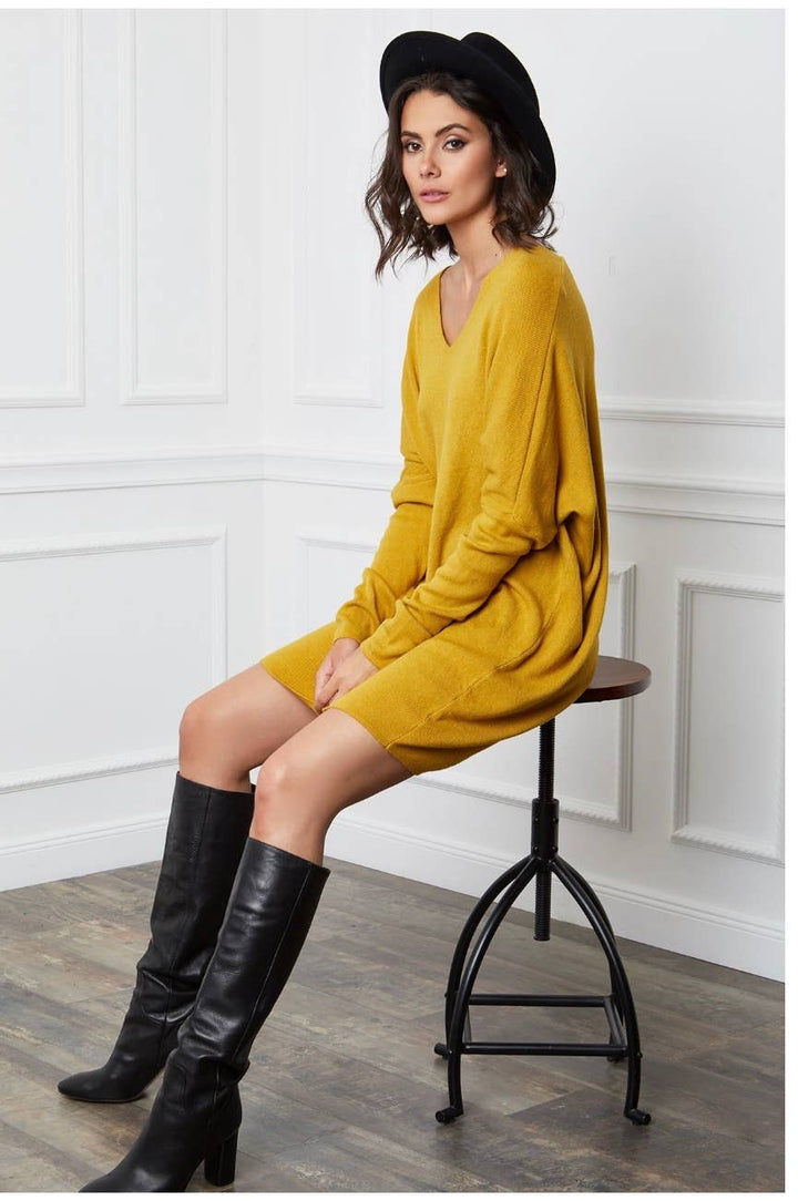 Woman in mustard yellow dress and black boots sitting on a stool against a white paneled wall.