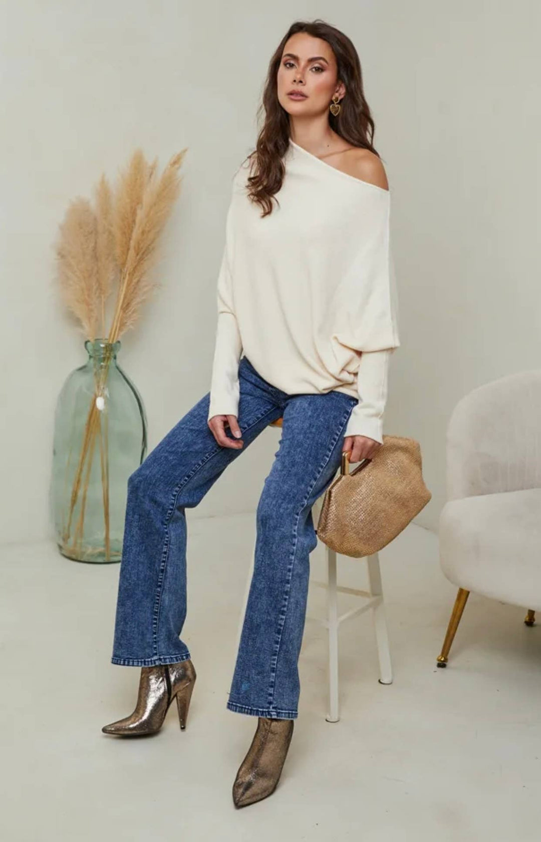 Woman in a white off-shoulder top and blue jeans sitting on a stool in a minimalistic room.