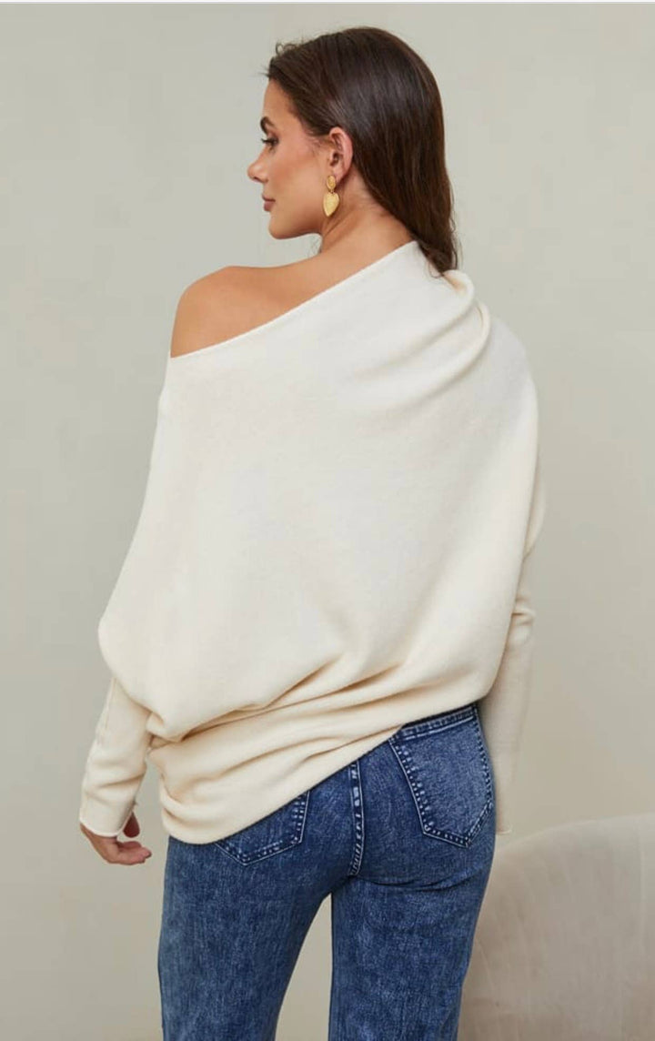 Woman wearing a cream off-shoulder sweater and blue jeans against a plain background
