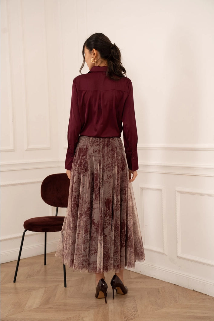 Woman wearing a burgundy blouse and patterned skirt standing in a room with a chair and white walls.