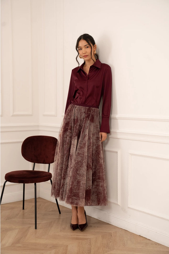 Woman wearing a burgundy shirt and pleated skirt standing against a white wall with a chair in the corner.