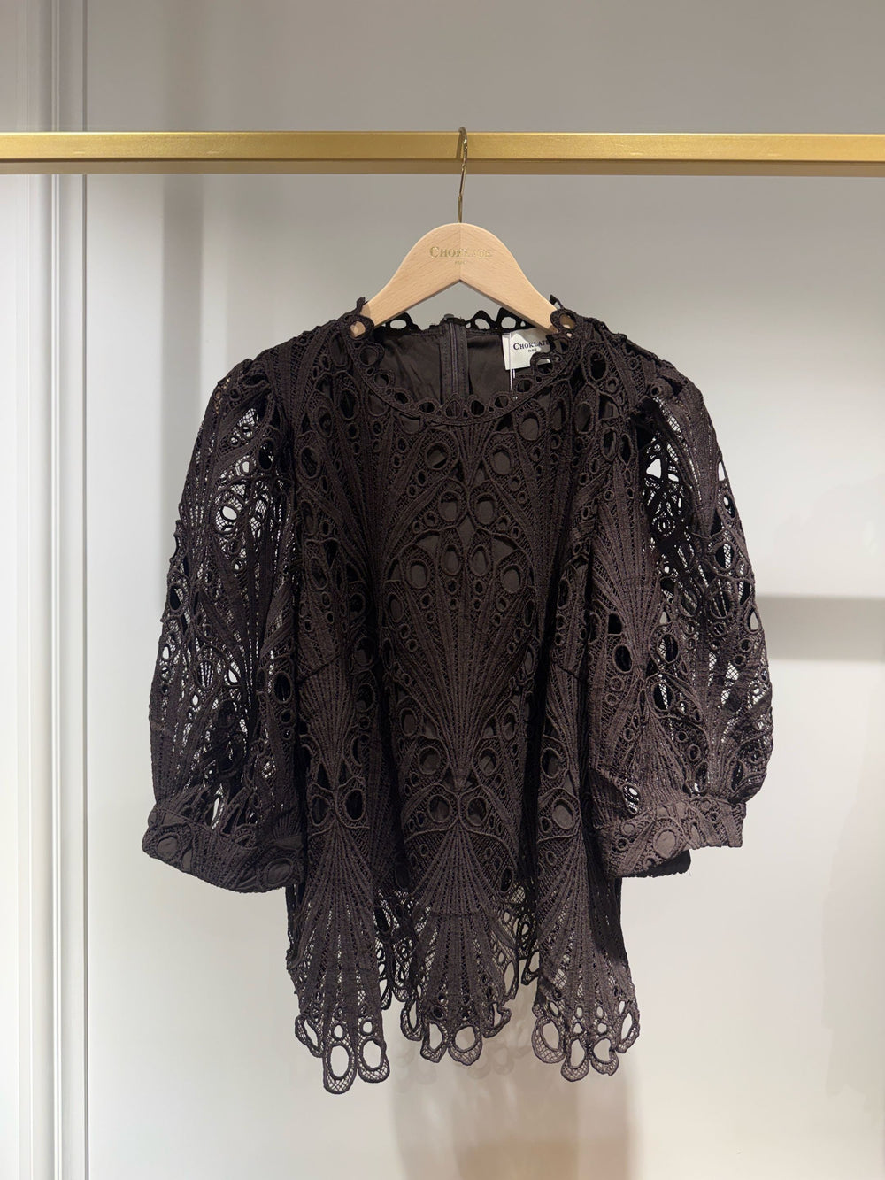 A dark brown eyelet crochet lace blouse with puff sleeves and a scalloped hem, hanging on a wooden hanger on a gold clothing rack.