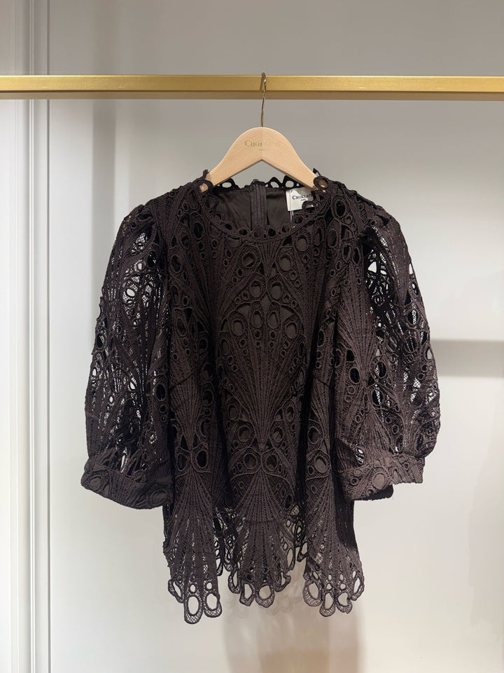 A dark brown eyelet crochet lace blouse with puff sleeves and a scalloped hem, hanging on a wooden hanger on a gold clothing rack.