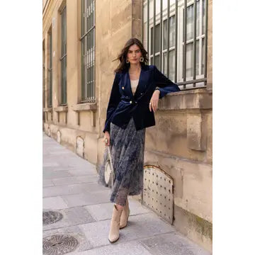 A woman stands on a city sidewalk beside a beige stone building, wearing a dark navy velvet blazer over a cream top and a gray patterned midi skirt, with beige ankle boots and a small white handbag.