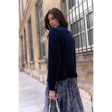 A woman wearing a dark navy velvet long-sleeve cardigan and a gray patterned skirt, standing on a city sidewalk next to a stone building.