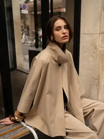 Woman wearing a beige trench coat sitting on a bench in an urban setting.