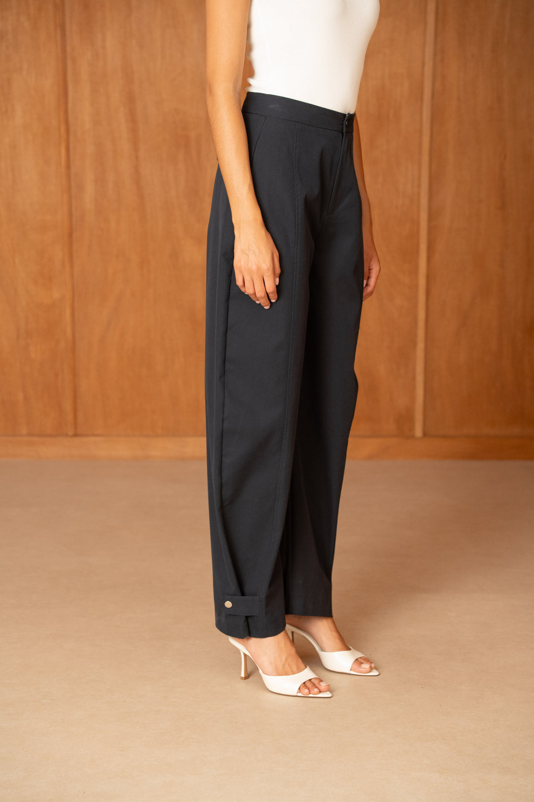 A model wearing black wide-leg cropped trousers with rolled cuffs and white heeled sandals, standing against a warm wood-paneled background.