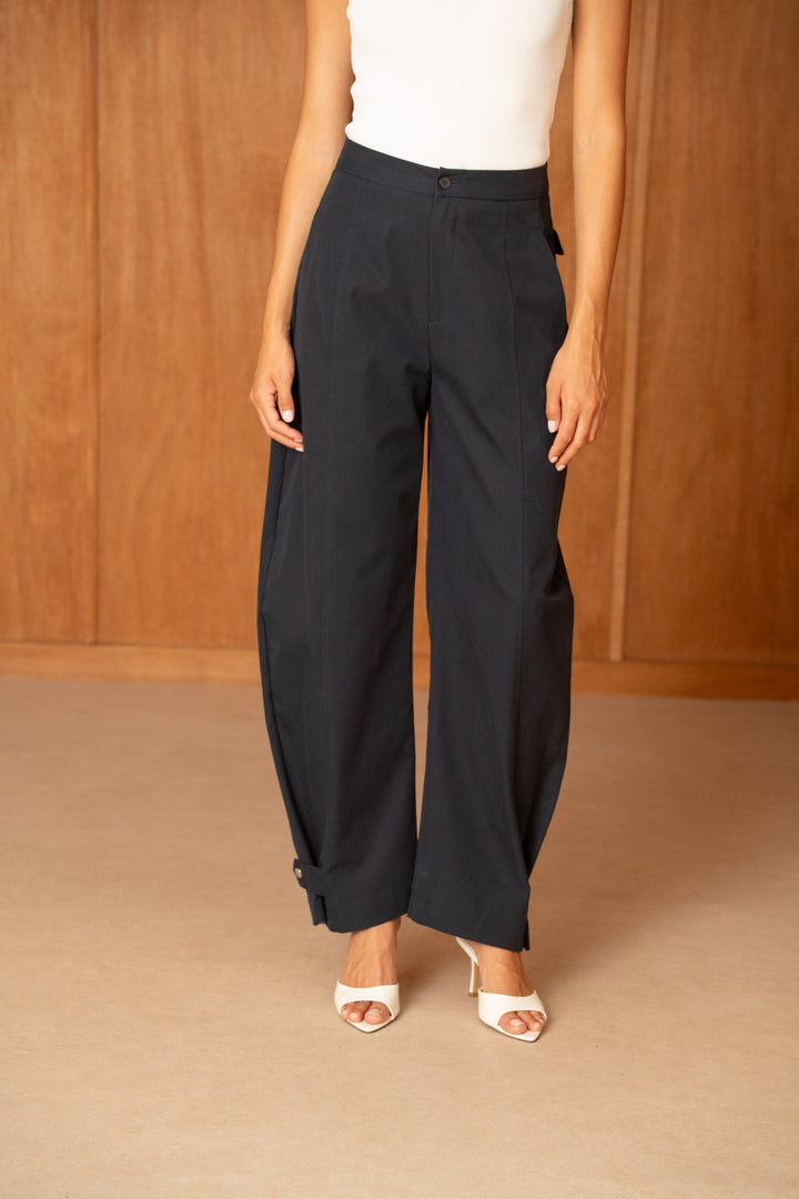 A model wearing black wide-leg cropped trousers with a white sleeveless top and white heeled sandals, standing against a warm wood-paneled background.
