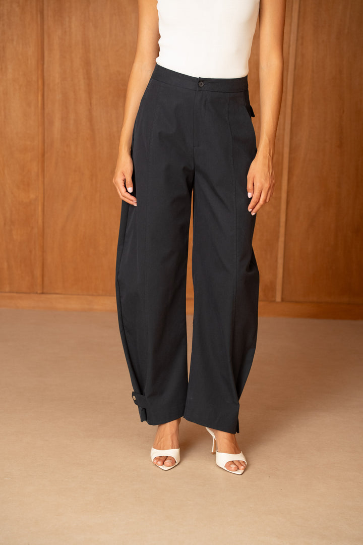 A model wearing black wide-leg cropped trousers with a white sleeveless top and white heeled sandals, standing against a warm wood-paneled background.