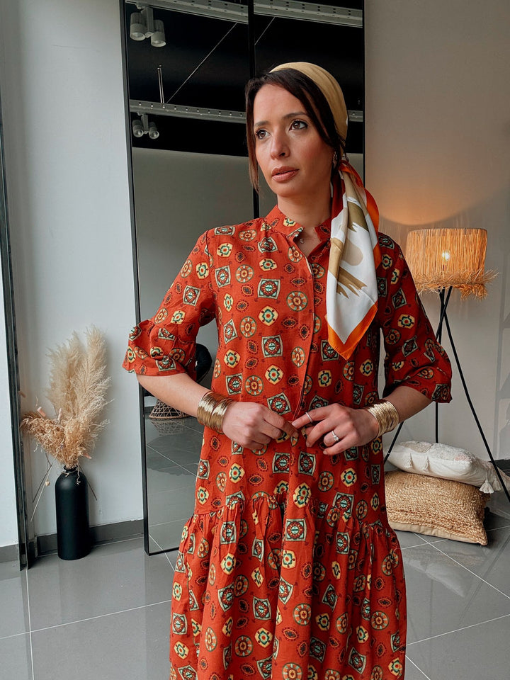Woman wearing a red patterned dress with a beige hat and scarf indoors.