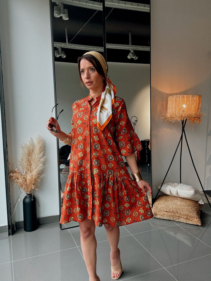 Woman in an orange patterned dress standing in a room with a lamp and decor.