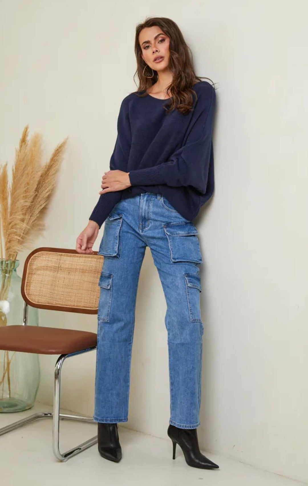 Woman wearing a navy sweater and blue jeans standing against a white wall.