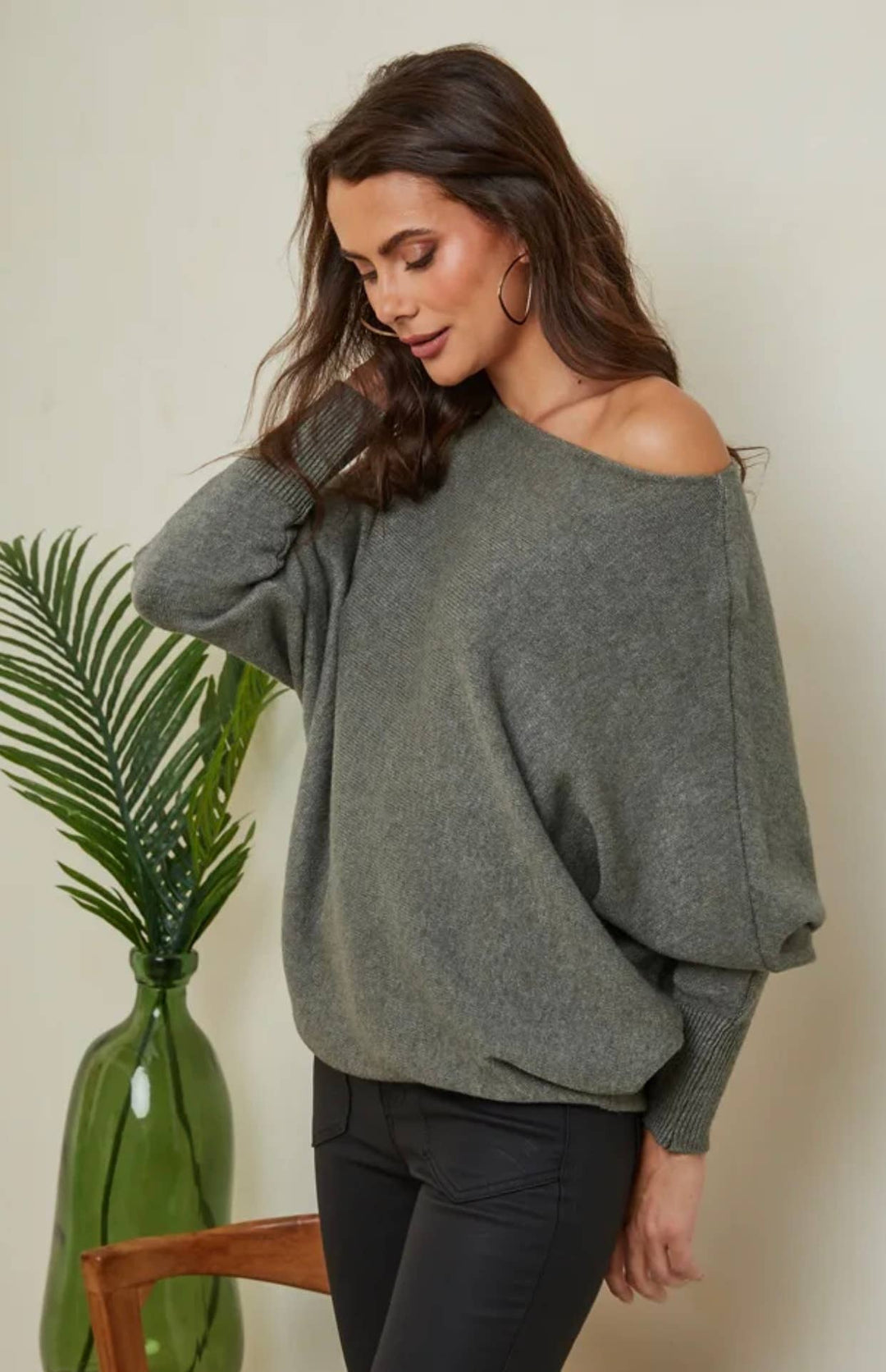 Woman wearing a gray off-shoulder sweater standing next to a plant against a beige wall.