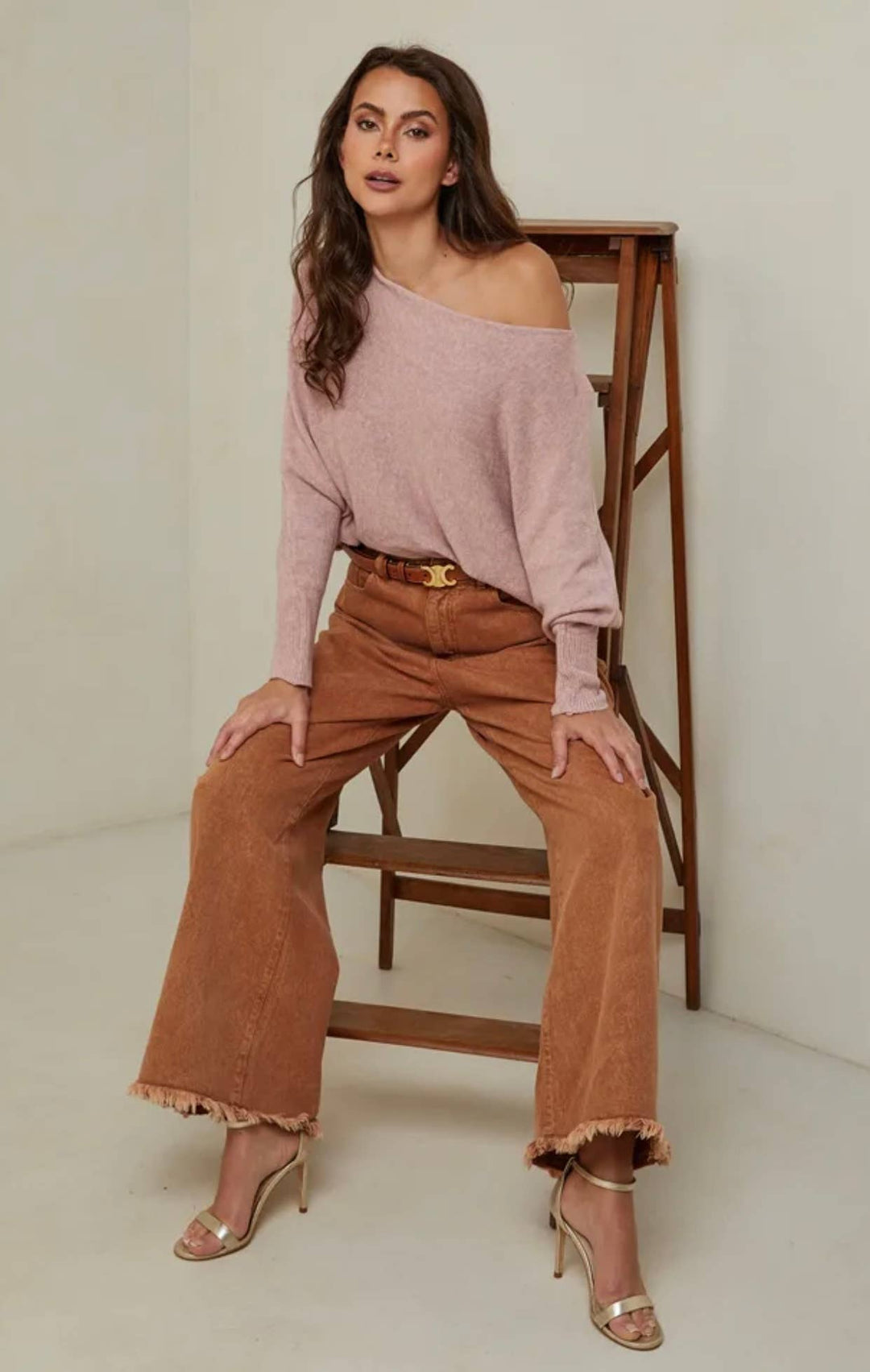 Woman wearing a pink off-shoulder top and brown wide-leg pants sitting on a wooden chair.
