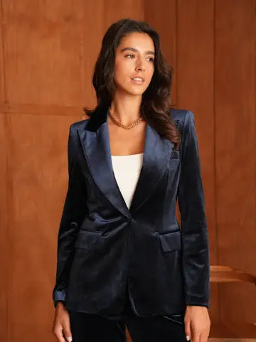 A woman wearing a dark navy blue velvet blazer over a white top, standing against a warm brown wood-paneled background.