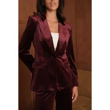 A woman wearing a deep burgundy satin blazer and matching trousers over a white top, shown from the mouth down against a warm brown indoor background.