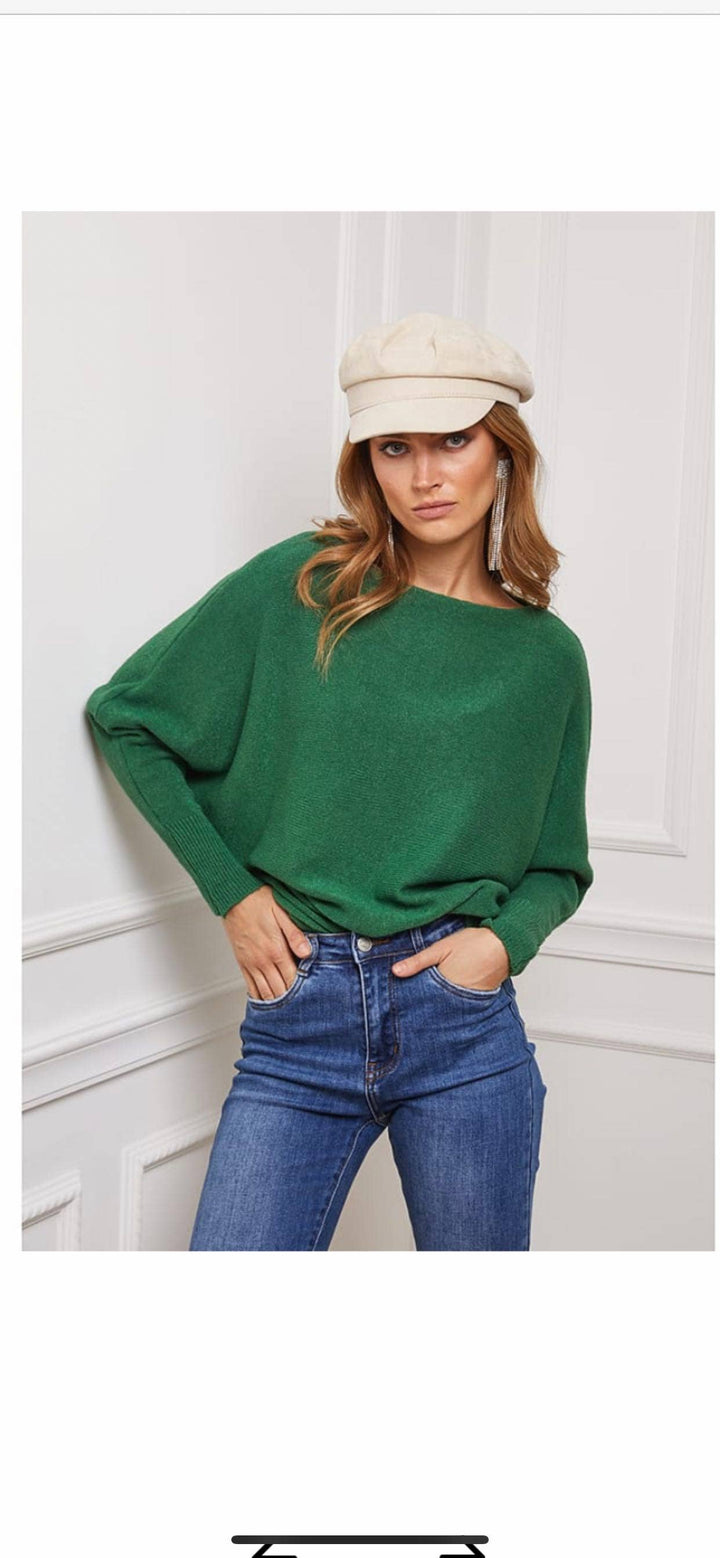 Woman wearing a green sweater, blue jeans, and a beige cap against a white wall.
