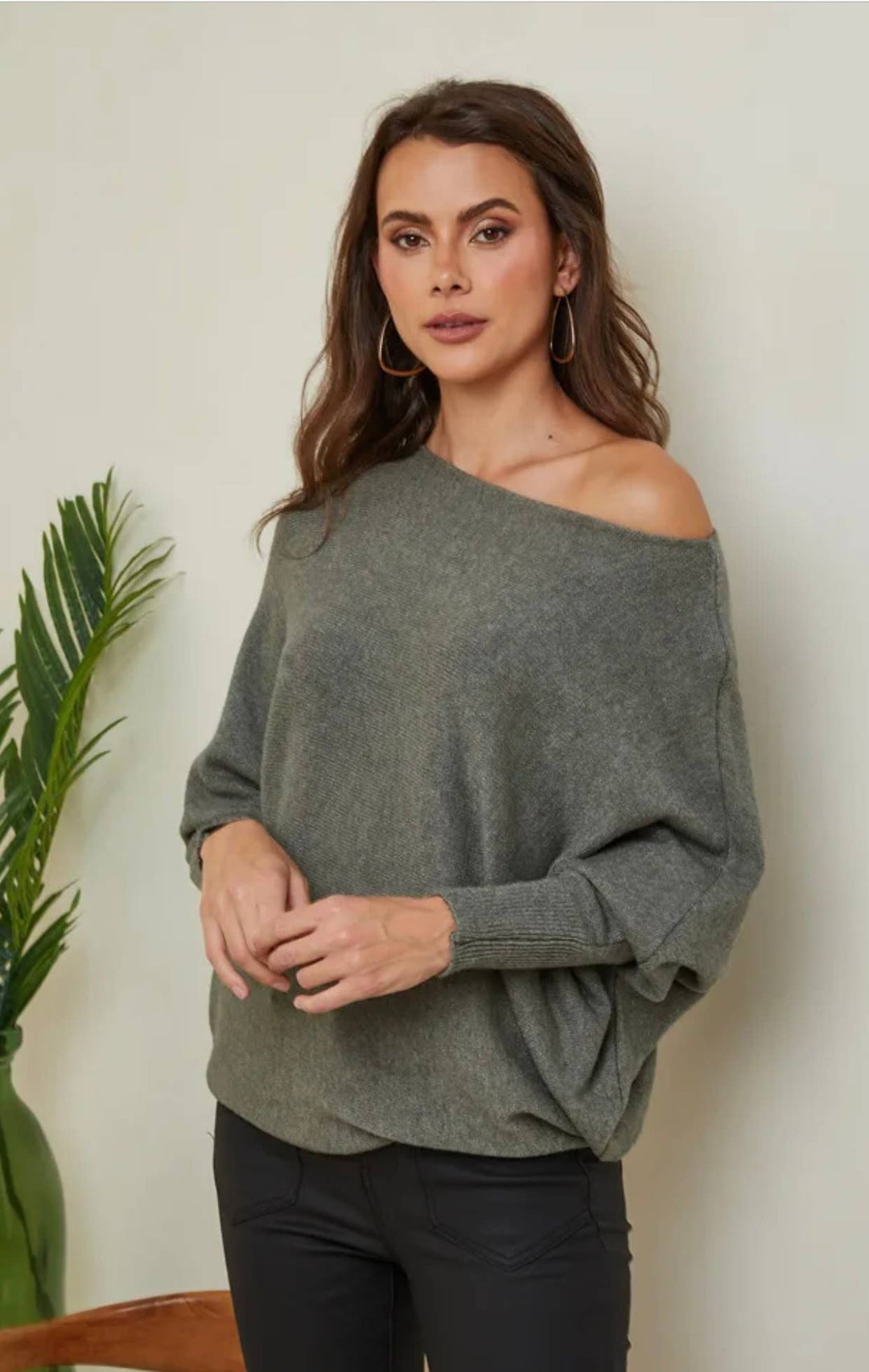 Woman wearing a green off-shoulder sweater against a white wall with a plant to the left.