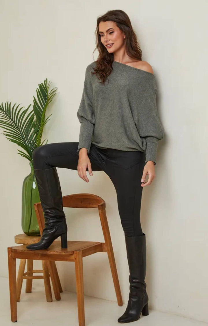 Woman wearing a gray off-shoulder sweater and black leather pants, standing next to a wooden chair with a plant.