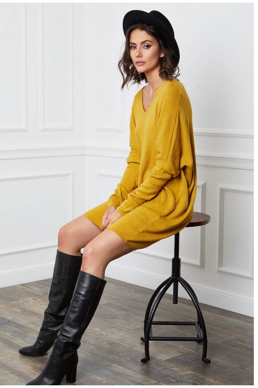Woman in mustard yellow dress and black boots sitting on a stool against a white paneled wall.