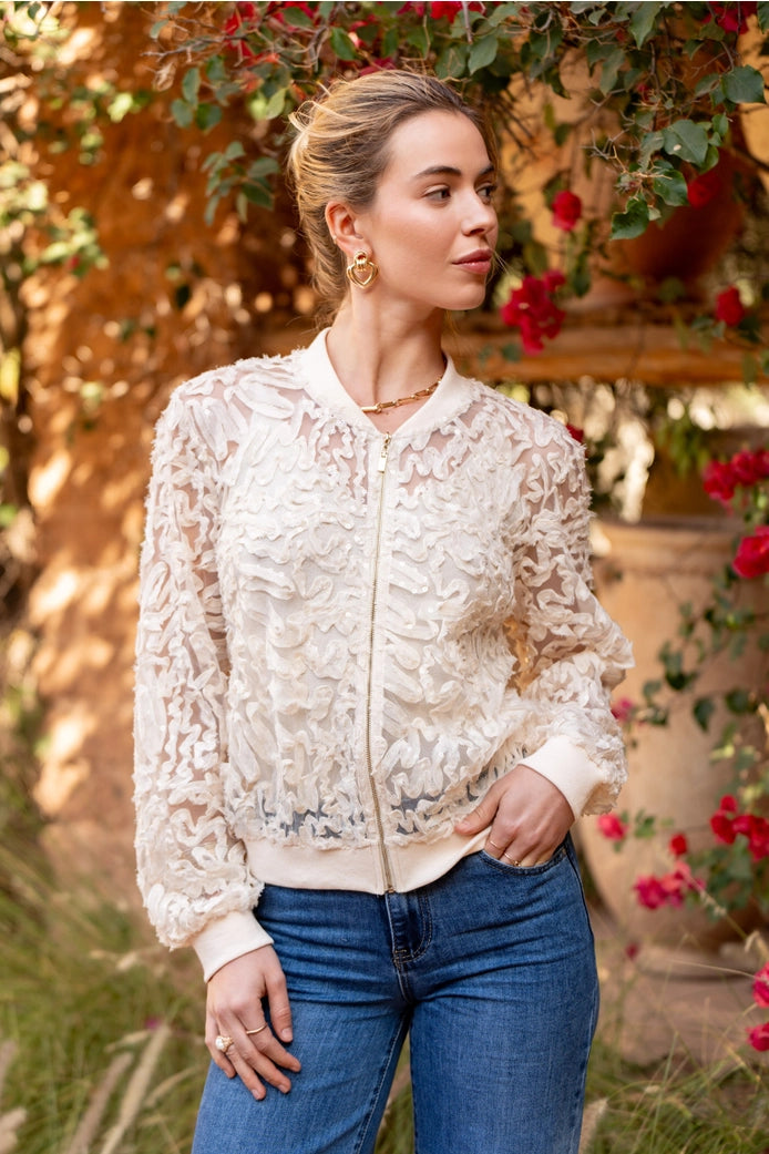 Woman wearing a white lace bomber jacket and blue jeans standing in a garden with flowers and a rustic wall.