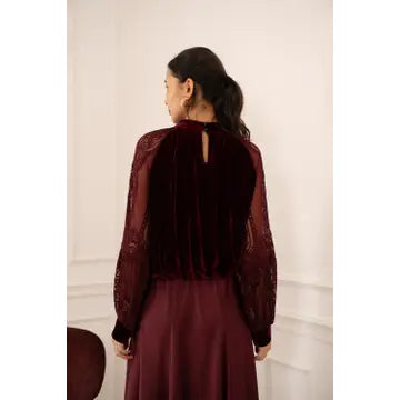 A woman viewed from behind wearing a burgundy long-sleeve dress with a velvet back panel and sheer lace sleeves, set against a light cream wall.