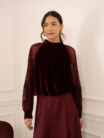 A woman wearing a dark burgundy velvet mock-neck top with sheer lace sleeves and a matching burgundy skirt, standing against a white paneled wall.