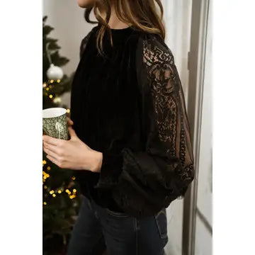 A woman wearing a black velvet long-sleeve blouse with sheer lace sleeves featuring a floral pattern, paired with dark jeans, holding a mug in an indoor setting.