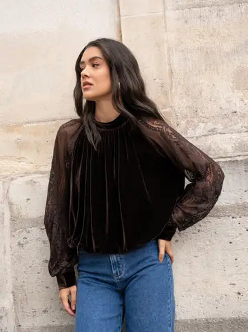 A woman wearing a dark brown velvet long-sleeve blouse with sheer lace sleeves, paired with blue jeans, standing against a textured stone wall.
