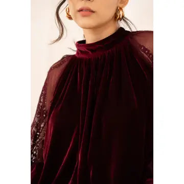 A close-up of a model wearing a deep burgundy velvet mock neck top with sheer embroidered sleeves, paired with gold hoop earrings.