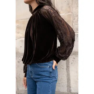 A woman wearing a dark brown velvet long-sleeve blouse with sheer lace sleeves and pleated detailing, paired with blue jeans, standing against a light stone wall.