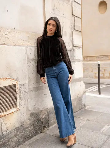 A woman stands on a city sidewalk against a textured stone wall, wearing a sheer black long-sleeve top with a velvet-like black bodice, blue wide-leg jeans, and tan heeled sandals.