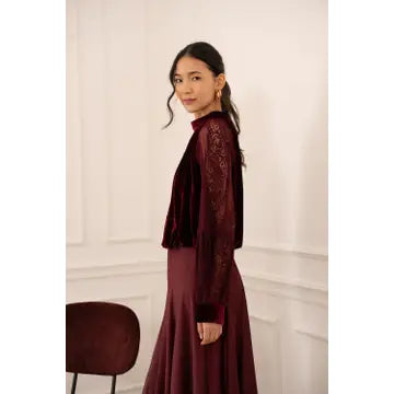 A woman wearing a long-sleeved burgundy velvet and lace dress, standing in a white paneled room.