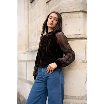 A woman wearing a dark brown sheer long-sleeve top with a velvet front and blue jeans, leaning against a light-colored stone wall.