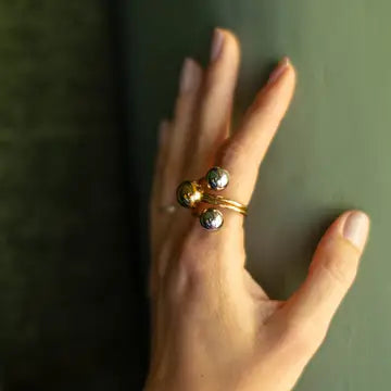 A close-up of a hand wearing a gold ring with three small metallic beads, set against a dark green background.