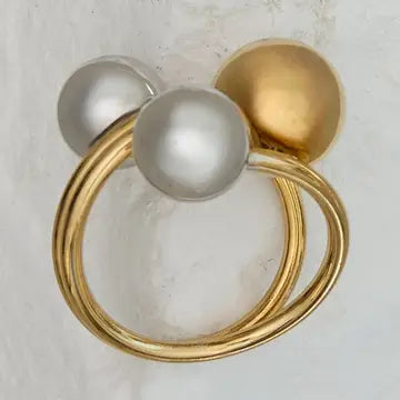 A gold ring featuring three large spherical beads, including two silver-toned pearls and one polished gold bead, set against a textured off-white background.