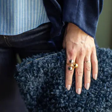 A close-up of a person's hand resting on a dark blue shaggy surface, wearing a statement ring with gold and black elements and a simple gold band.