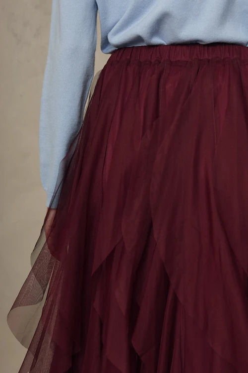 A close-up of a person wearing a long, layered tulle skirt in deep burgundy with a light blue top, shown from the waist down against a neutral beige background.