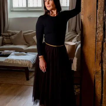 A woman wearing a black long-sleeve top and a long black tulle skirt with a thin gold waistband, standing in a cozy living room next to a rustic wooden pillar.