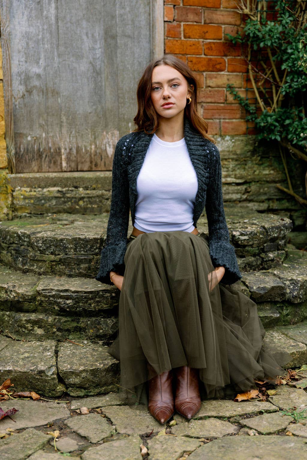 Woman standing on stone steps wearing a green cardigan, white top, and brown skirt.